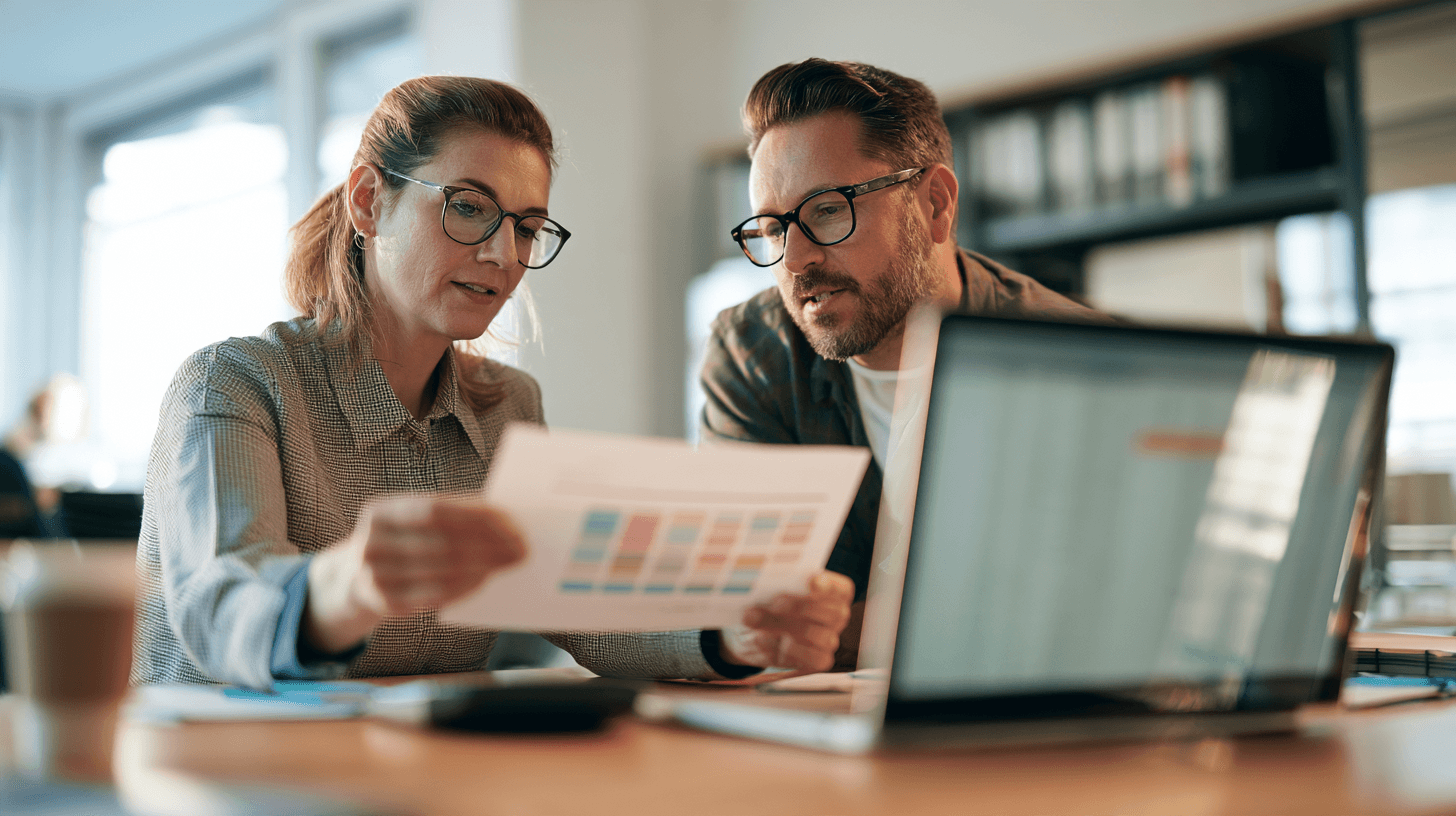 HR-Managerin reicht ihrem Kollegen eine ausgedruckte Merit-Matrix für die jährliche Gehaltsrunde am Schreibtisch